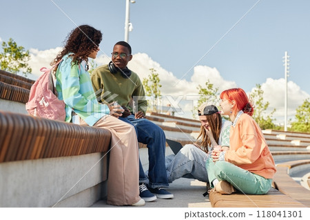 Side view of diverse group of teenage students engaged in animated conversation sitting on amphitheater bench while hanging out together outdoors on sunny day, copy space 118041301