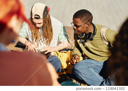 Teenage African American college student in glasses curiously looking at friends laptop, while classmate surfing net doing homework on bench outdoors enjoying sunny weather after classes Teenage African American college student in glasses curiously looking at friends laptop, while classmate surfing net doing homework on bench outdoors enjoying sunny weather after classes 118041302