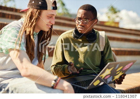 Medium shot of Black teenage boy with frowning face looking intently on male friend explaining something, while surfing net together using laptop on bench in park 118041307