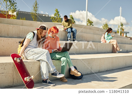 Smiling teenage male skateboarder and redhead girl in bright outfit having fun while using laptop together on concrete seat of amphitheater in park Smiling teenage male skateboarder and redhead girl in bright outfit having fun while using laptop together on concrete seat of amphitheater in park 118041310