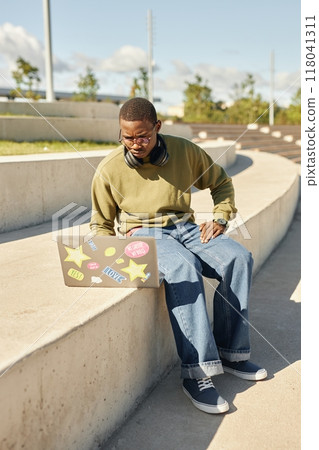 Vertical shot of concentrated Black teenage boy with frowning face using laptop on concrete seat of amphitheater in park, while studying after classes outdoors on sunny day Vertical shot of concentrated Black teenage boy with frowning face using laptop on concrete seat of amphitheater in park, while studying after classes outdoors on sunny day 118041311