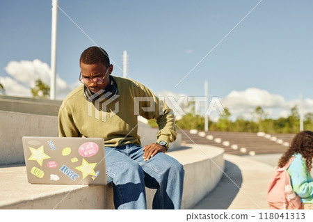 Concentrated Black teenage boy with frowning face using laptop on concrete seat of amphitheater in park while studying after classes outdoors on sunny day, copy space 118041313