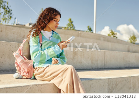 Side view of multiethnic curly girl with schoolbag dressed in pastel clothes using phone surfing net while sitting on concrete bench in park, copy space Side view of multiethnic curly girl with schoolbag dressed in pastel clothes using phone surfing net while sitting on concrete bench in park, copy space 118041318