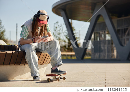 Teenage skateboarder in headphones on phone text messaging while sitting on park bench during sunny early autumn day, copy space Teenage skateboarder in headphones on phone text messaging while sitting on park bench during sunny early autumn day, copy space 118041325