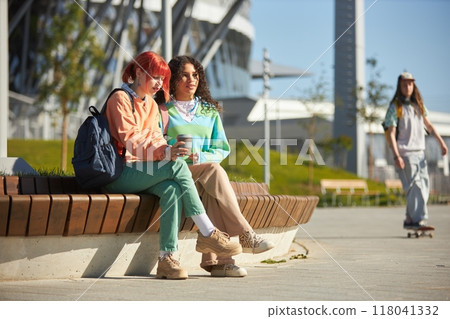Two teenage girls of diverse ethnicity dressed in bright vibrant clothes sitting together on wooden park bench during break enjoying sunny day on college campus, copy space Two teenage girls of diverse ethnicity dressed in bright vibrant clothes sitting together on wooden park bench during break enjoying sunny day on college campus, copy space 118041332