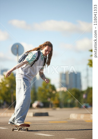 Vertical shot of cool dreadlocked teenage boy with backpack riding skateboard down sunlit city street to school captured against early autumn sky, copy space Vertical shot of cool dreadlocked teenage boy with backpack riding skateboard down sunlit city street to school captured against early autumn sky, copy space 118041333