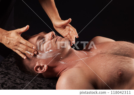 A man is getting a facial treatment at a spa. He is lying on a dark surface with his eyes closed, and the masseur is applying cream to his face with gentle hands. Facial massage concept A man is getting a facial treatment at a spa. He is lying on a dark surface with his eyes closed, and the masseur is applying cream to his face with gentle hands. Facial massage concept 118042311