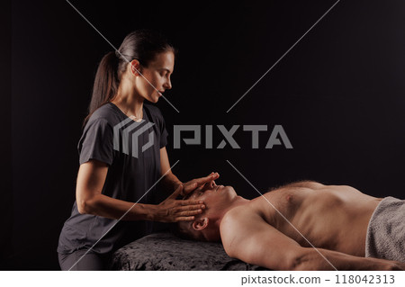 Female masseur doing facial massage to man lying on massage table.on dark uniform background, facial massage concept 118042313