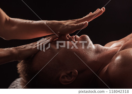 Close-up of a man receiving a facial massage against a dark background, The hands of the massage therapist are gently pressing on his forehead and temples. 118042318
