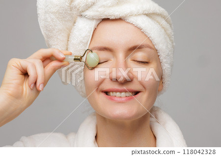Woman with towel wrapped around head using jade roller on face smiling. Roller massaging eyes demonstrating skin care routine. Woman with towel wrapped around head using jade roller on face smiling. Roller massaging eyes demonstrating skin care routine. 118042328