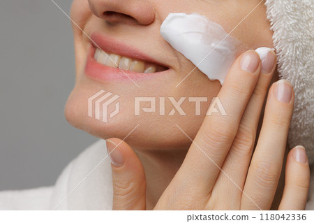A woman applying cream to her face, smiling gently. She has a towel wrapped around her head and is in a relaxed setting, suggesting a skincare routine or spa treatment. 118042336