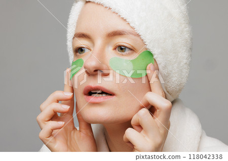 A woman with a towel on her head and a robe smiles as she applies green gel patches under her eyes facial cosmetology concept 118042338