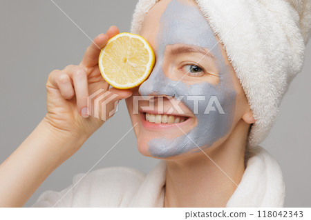 A woman with a mask on her face holding a lemon wedge in front of her mouth. She is wearing a white robe and a towel on her head. Beauty and Skin care concept 118042343