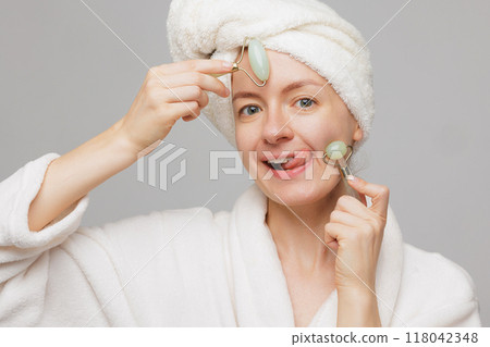 woman with towel on head and robe holding jade rollers on cheeks sticking out tongue, wrinkle smoothing concept 118042348