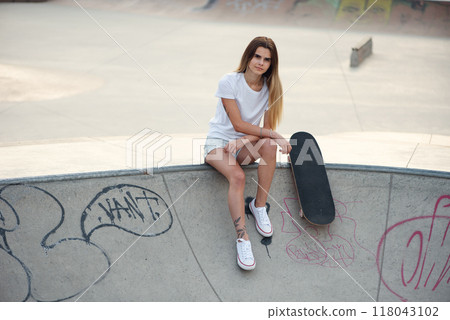 Young attractive girl in a white t-shirt and sneakers in modern skatepark with skateboard. Young attractive girl in a white t-shirt and sneakers in modern skatepark with skateboard. 118043102