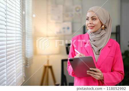 A woman in a pink jacket is holding a tablet and writing on a notepad A woman in a pink jacket is holding a tablet and writing on a notepad 118052992
