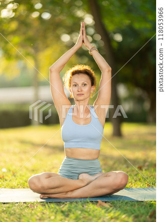 Portrait of woman sitting on gym mat and doing yoga meditation in park 118053966