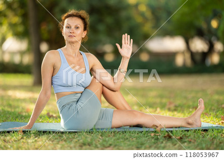 Sporty woman performing seated twisting asana Matsyendrasana on mat in in summer park during daily yoga session 118053967