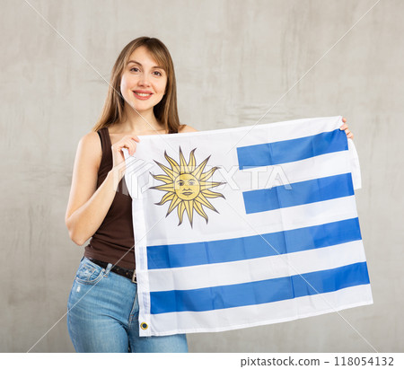 Happy cheerful young woman waving national flag of Uruguay while looking at camera at gray wall background 118054132