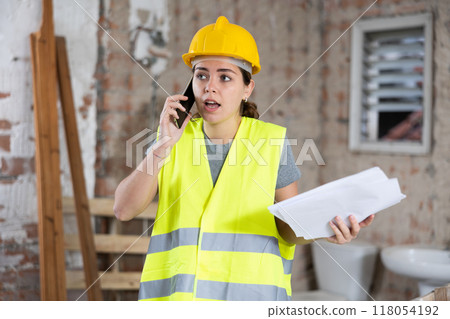 Young woman civil engineer with papers talking on phone at construction site Young woman civil engineer with papers talking on phone at construction site 118054192