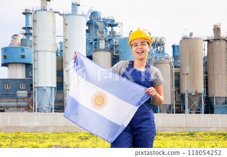 Young female engineer in helmet waving state flag of Argentina while standing in front of big tanks at chemical plant 118054252