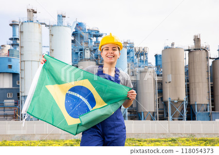 Young female engineer in helmet waving state flag of Brazil while standing in front of big tanks at chemical plant 118054373