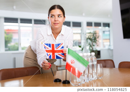 A woman in a white blouse with friendly smile arrangs flags of Britain and Iran for meeting. A woman in a white blouse with friendly smile arrangs flags of Britain and Iran for meeting. 118054825