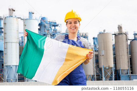 Young female engineer in helmet waving state flag of Ireland while standing in front of big tanks at chemical plant 118055116