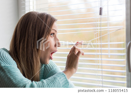 Surprised woman peeking through window blinds on a bright day, capturing unexpected moments in her home 118055415