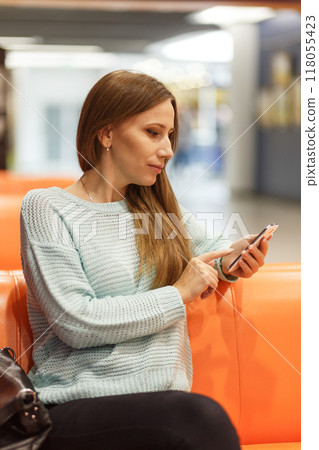 Young woman sitting on an orange bench using a smartphone in a modern, well-lit indoor space during daytime Young woman sitting on an orange bench using a smartphone in a modern, well-lit indoor space during daytime 118055423