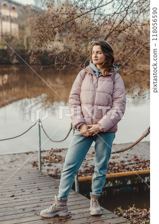 A young woman standing at the shore looking at the river in autumn sunny day. Street view, copy space for text, travel photo. Happy tourist woman on the bank of the river in autumn in warm clothes A young woman standing at the shore looking at the river in autumn sunny day. Street view, copy space for text, travel photo. Happy tourist woman on the bank of the river in autumn in warm clothes 118056590