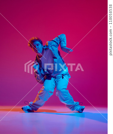 Dance club member. Young energetic man in casual clothes in motion, dance hip hop against pink background in neon light Dance club member. Young energetic man in casual clothes in motion, dance hip hop against pink background in neon light 118058538