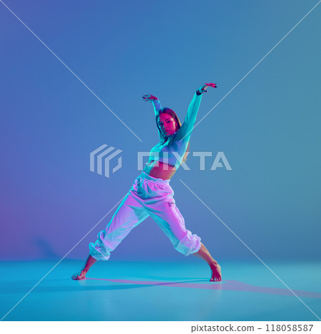 Youthful energy, dynamic hip hop choreography. Young talented and artistic girl performing against gradient blue background in neon light Youthful energy, dynamic hip hop choreography. Young talented and artistic girl performing against gradient blue background in neon light 118058587