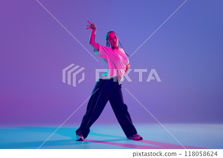 Young beautiful girl in stylish clothes performing hip hop with passion and energy against gradient purple background in neon light Young beautiful girl in stylish clothes performing hip hop with passion and energy against gradient purple background in neon light 118058624