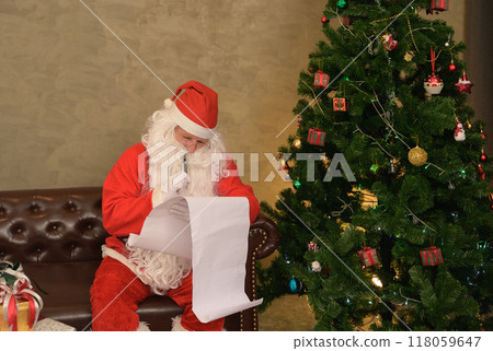Santa Claus checking long list for Christmas presents near decorated christmas tree in living room at home. Christmas and New Year concept. Santa Claus checking long list for Christmas presents near decorated christmas tree in living room at home. Christmas and New Year concept. 118059647