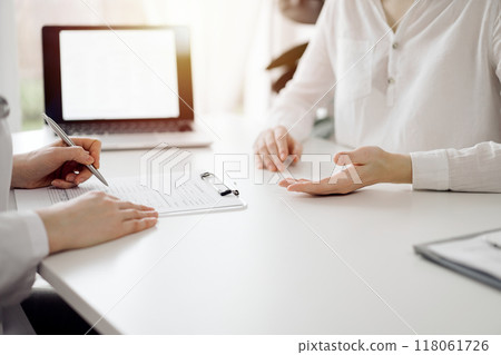 Doctor and patient sitting and discussing something near each other at the white desk in clinic. Female physician is listening filling up a records form. Medicine concept 118061726