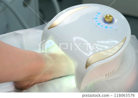 Woman is drying base coal shellac on nails on toes in UV lamp in beauty salon. Woman is drying base coal shellac on nails on toes in UV lamp in beauty salon. 118063576