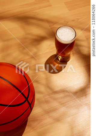 Pint glass of beer with foam sits on wooden floor. Sunlight casts shadow of basketball hoop on court, floor, while basketball rests nearby 118064266