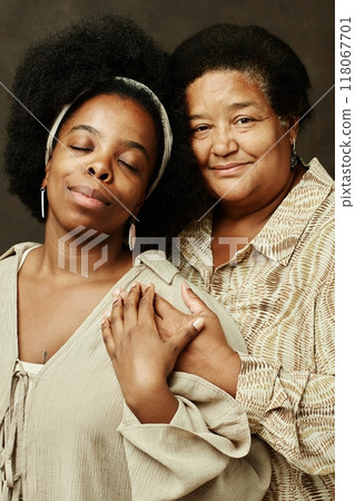 Vertical portrait shot of smiling elderly African American woman looking at camera gently hugging young daughter with eyes closed, while wearing beige clothes and posing on brown background in studio 118067701