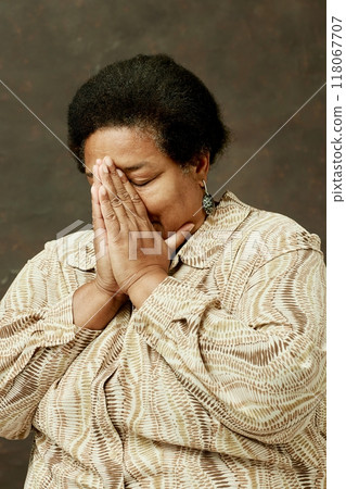 Medium shot of worried elderly African American woman in beige clothes covering face with folded hands, while praying with eyes closed on brown background in studio 118067707