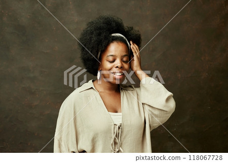 Chest up shot of graceful smiling Black woman with eyes closed touching natural curly hair while posing in beige clothes wearing no makeup on brown background in studio, copy space 118067728