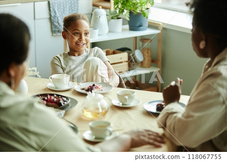 Young smiling Black girl sitting at table talking to family over cup of tea with delicious dessert on little plate at sunlit kitchen, copy space 118067755