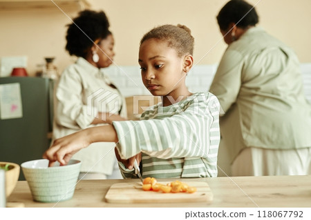 Medium shot of little Black girl in casual clothes taking vegetable from bowl cutting tomatoes while cooking fresh salad for healthy dinner at cozy beige kitchen, copy space 118067792