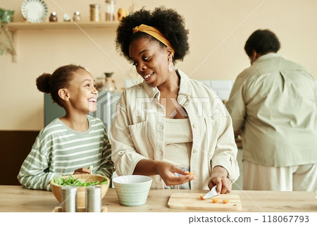 Medium shot of African American young mother and happy little daughter talking and smiling while making healthy salad together at cozy beige kitchen 118067793