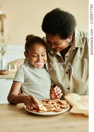 Vertical medium shot of smiling young Black girl in gentle hugs of happy grandmother serving fresh homemade biscuits on plate while cooking together at kitchen 118067794