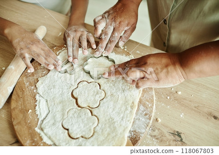 Close up on hands of African American woman and young girl cutting flower shaped cookies from rolled out dough with metal cookie cutters, while making homemade biscuits on wooden kitchen counter 118067803