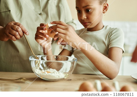 Cropped shot of concentrated African American little girl carefully cracking egg in bowl at wooden counter, learning how to cook pancakes together with granny at beige kitchen Cropped shot of concentrated African American little girl carefully cracking egg in bowl at wooden counter, learning how to cook pancakes together with granny at beige kitchen 118067811