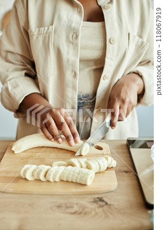 Vertical shot of hands of unrecognizable Black woman in nude clothes cutting bananas for healthy breakfast at beige kitchen counter 118067919