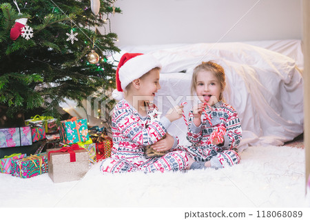 Children in festive pajamas eating Christmas candy canes from jar of holiday treats beside decorated Christmas tree during the festive season. Joyful siblings sit on cozy rug, enjoying festive sweets 118068089