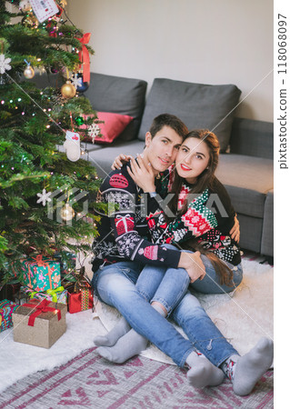 Portrait of Happy Caucasian couple in festive Christmas jumpers hugging while sitting under decorated Christmas tree, sharing joyful moment during holiday season at home. Romantic Holidays together. Portrait of Happy Caucasian couple in festive Christmas jumpers hugging while sitting under decorated Christmas tree, sharing joyful moment during holiday season at home. Romantic Holidays together. 118068097